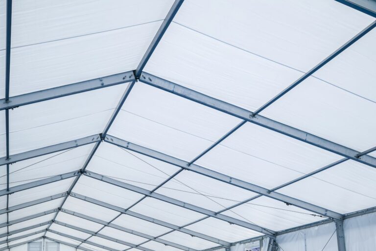 Structură metalică de acoperiș acoperită cu material textil alb, văzută din interiorul unui cort industrial sau al unei hale temporare.