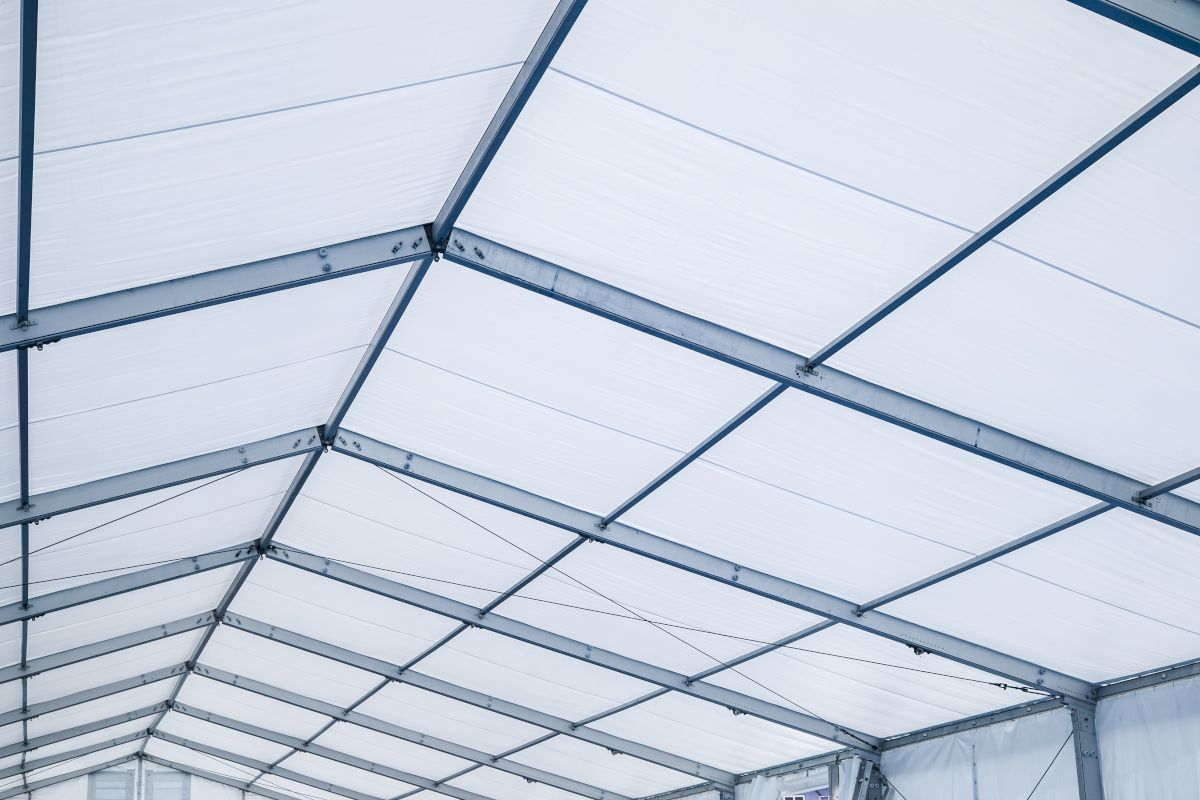 Structură metalică de acoperiș acoperită cu material textil alb, văzută din interiorul unui cort industrial sau al unei hale temporare.